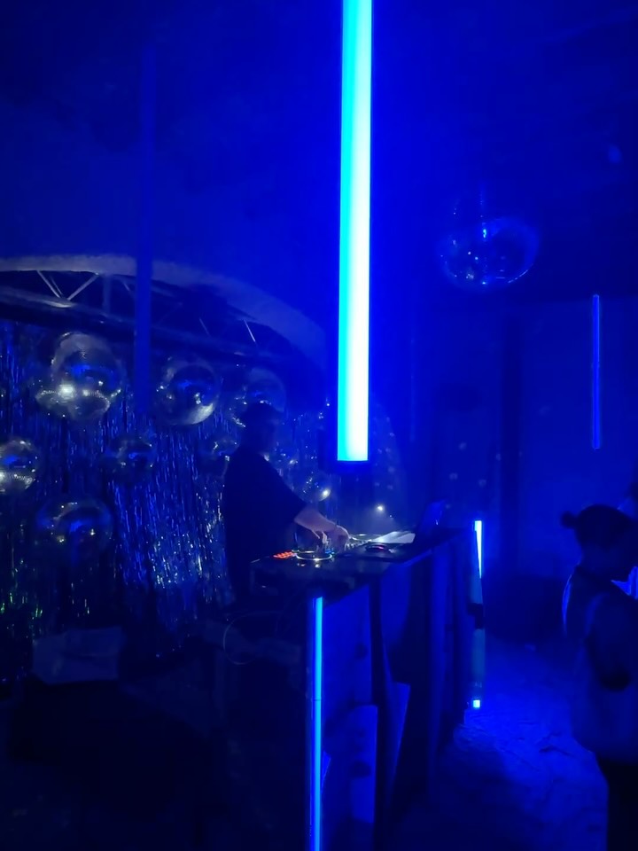 Andrew Mackenzie performing behind the decks under blue tube lights and disco balls